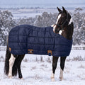 A dark brown and white pinto horse standing in a snowy field, wearing a navy blue quilted horse blanket with brown leather accents NAVY1